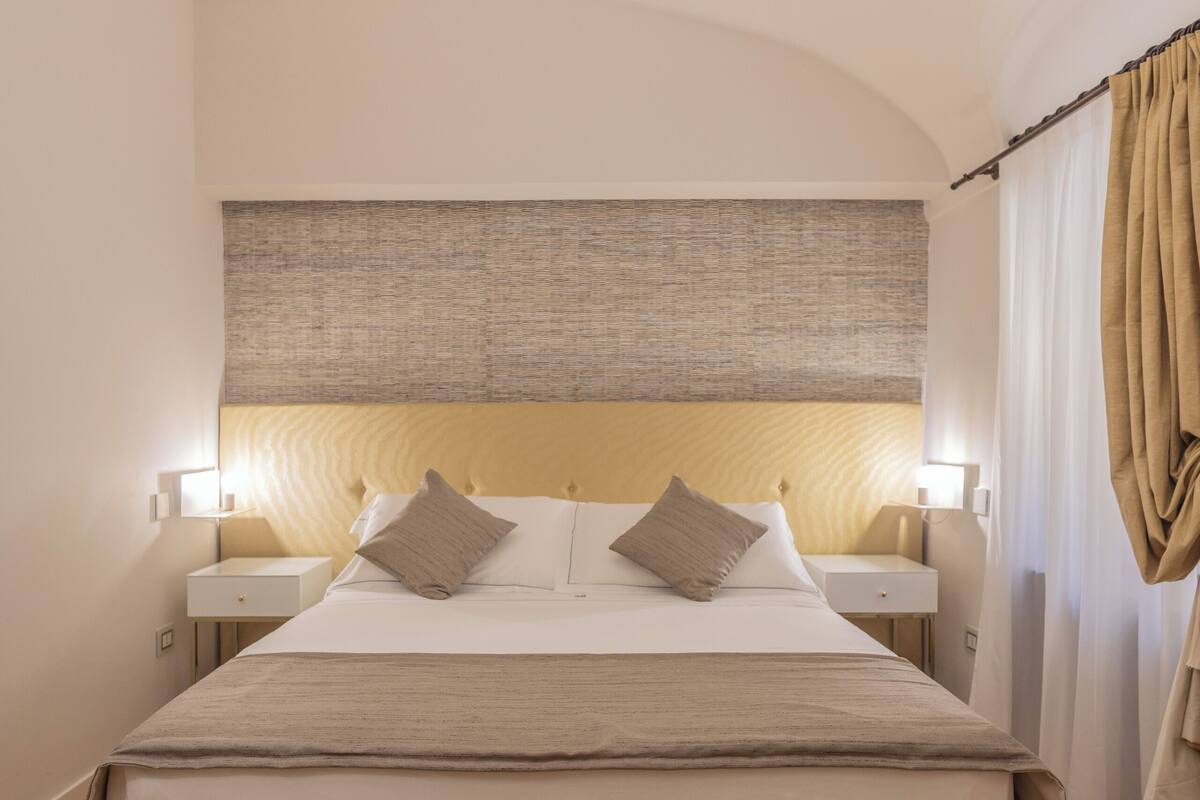 Un dormitorio con una cama grande, dos mesitas de noche con lámparas, un cabecero con panel texturizado y piso de madera.