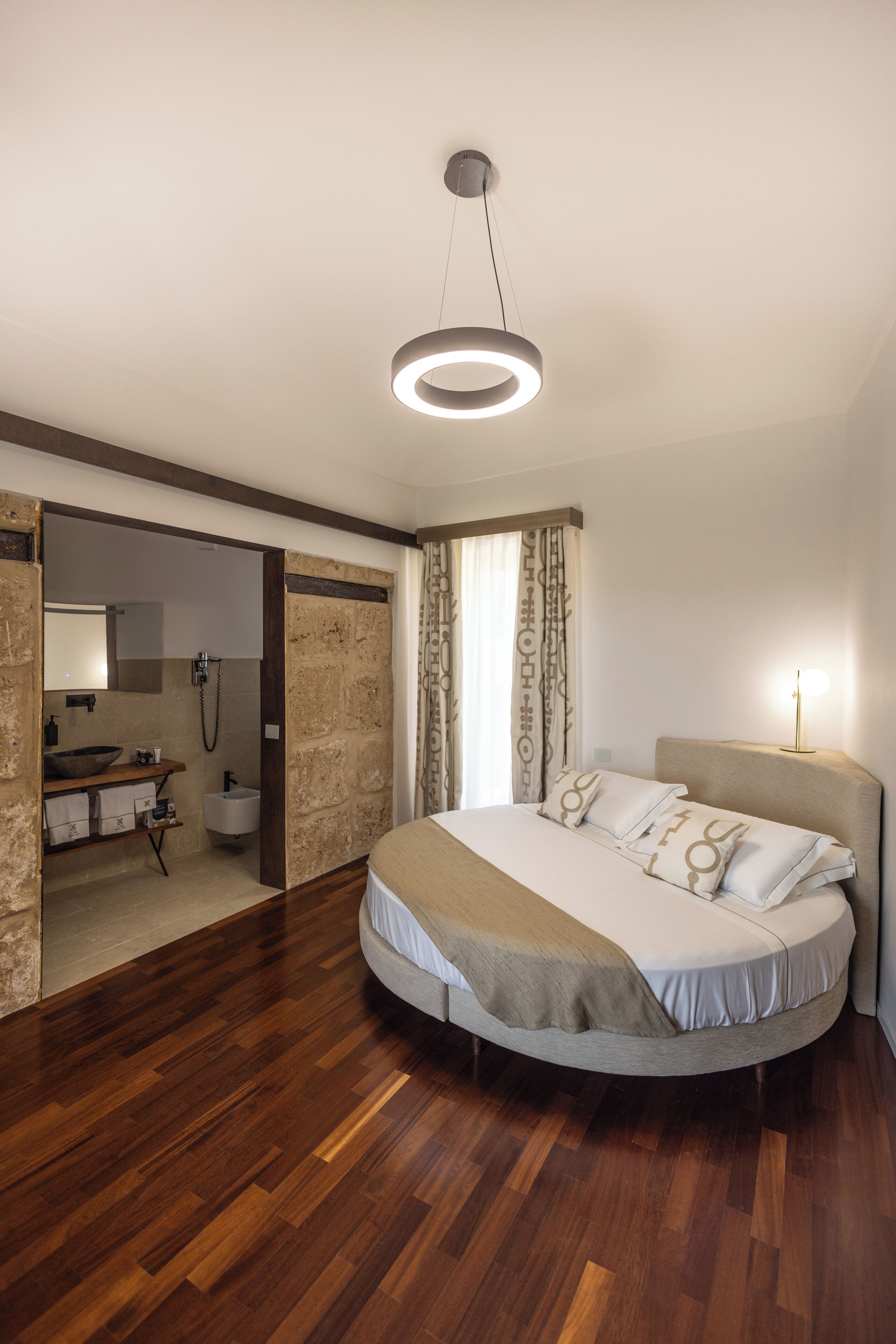 Un dormitorio moderno con una cama grande, suelo de madera, un armario empotrado, un baño con lavamanos y espejo, y una lámpara de techo circular.