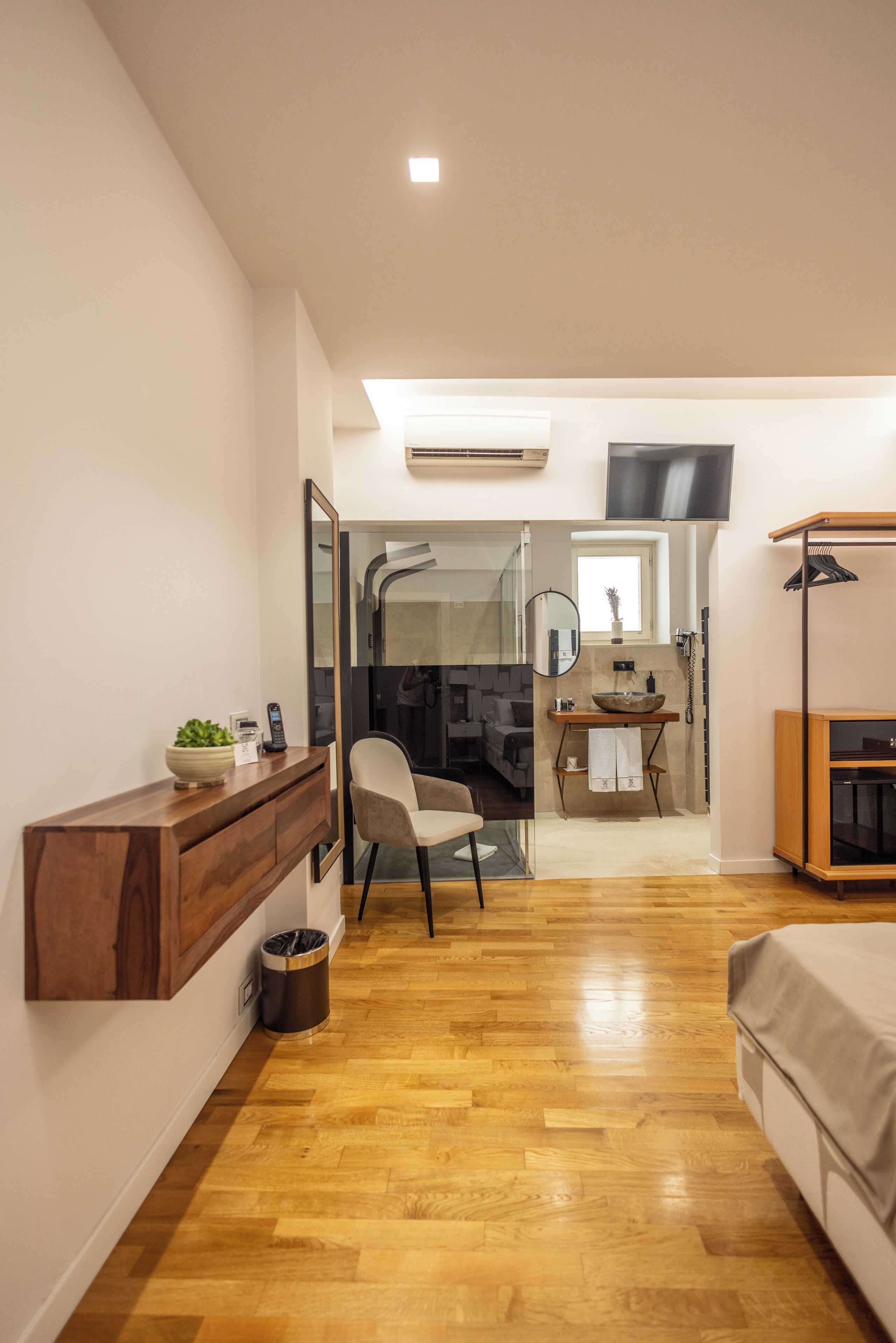 A modern hotel room with a wooden floor, a desk with a mirror, a chair, a bed, and a TV mounted on the wall.