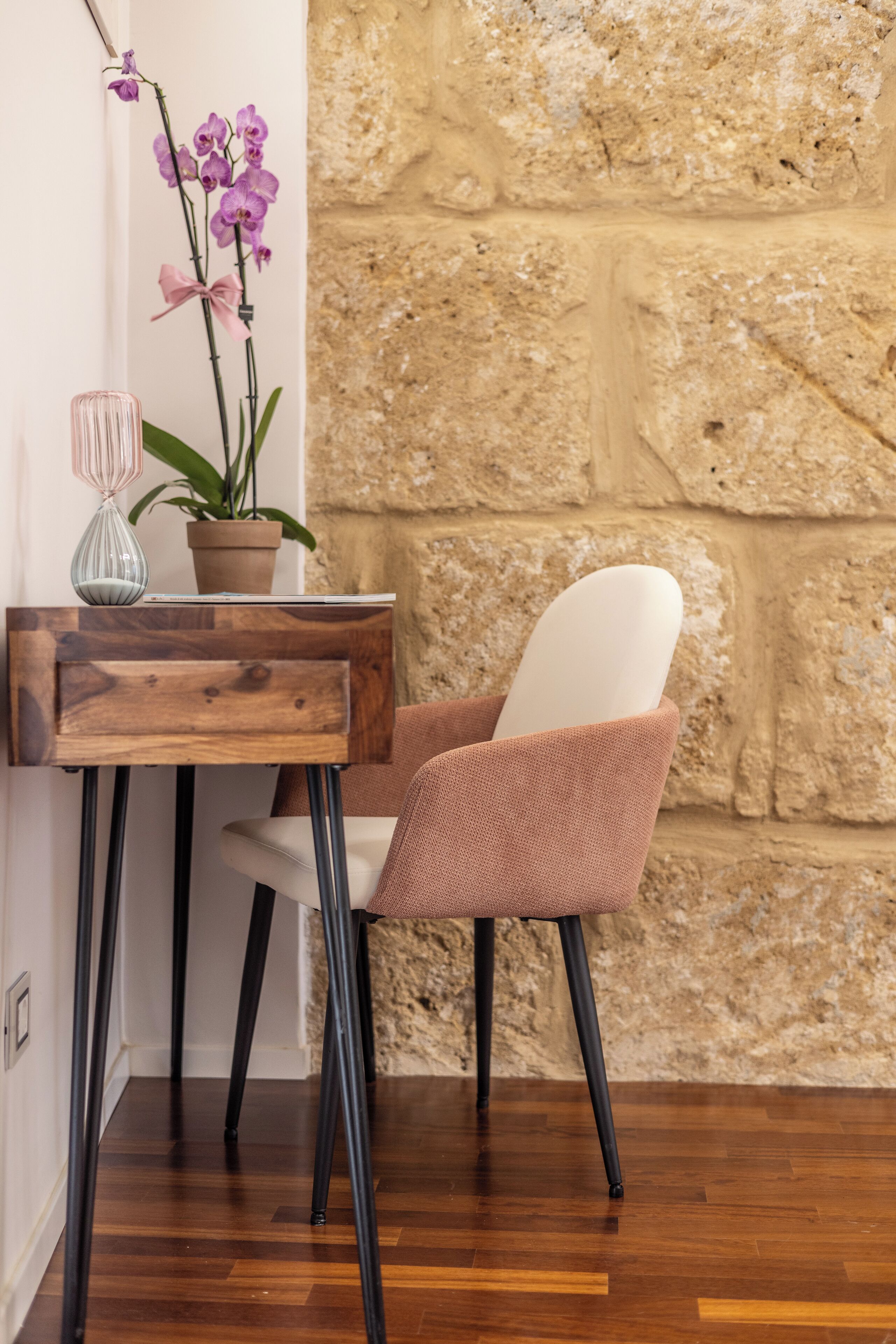 A wooden table with a vase and a potted plant, a chair with a cushion, and a stone wall.