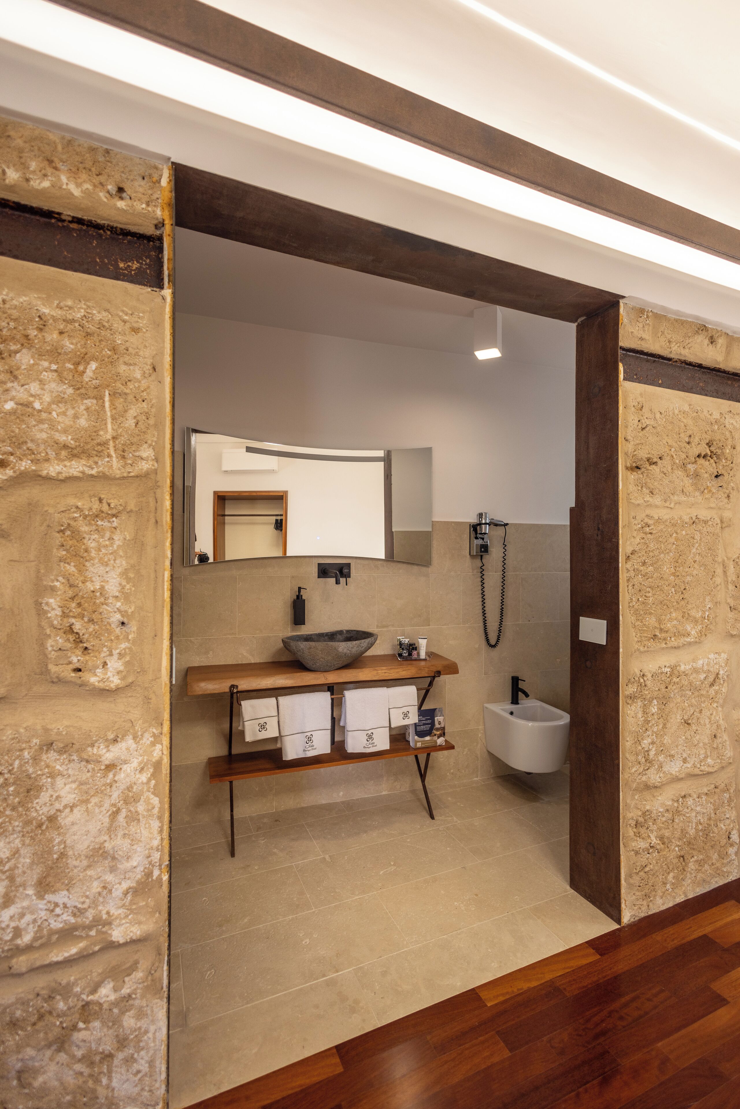 A bathroom with a stone wall, wooden floor, a sink with a basin, a mirror, a bidet, and a towel rack.