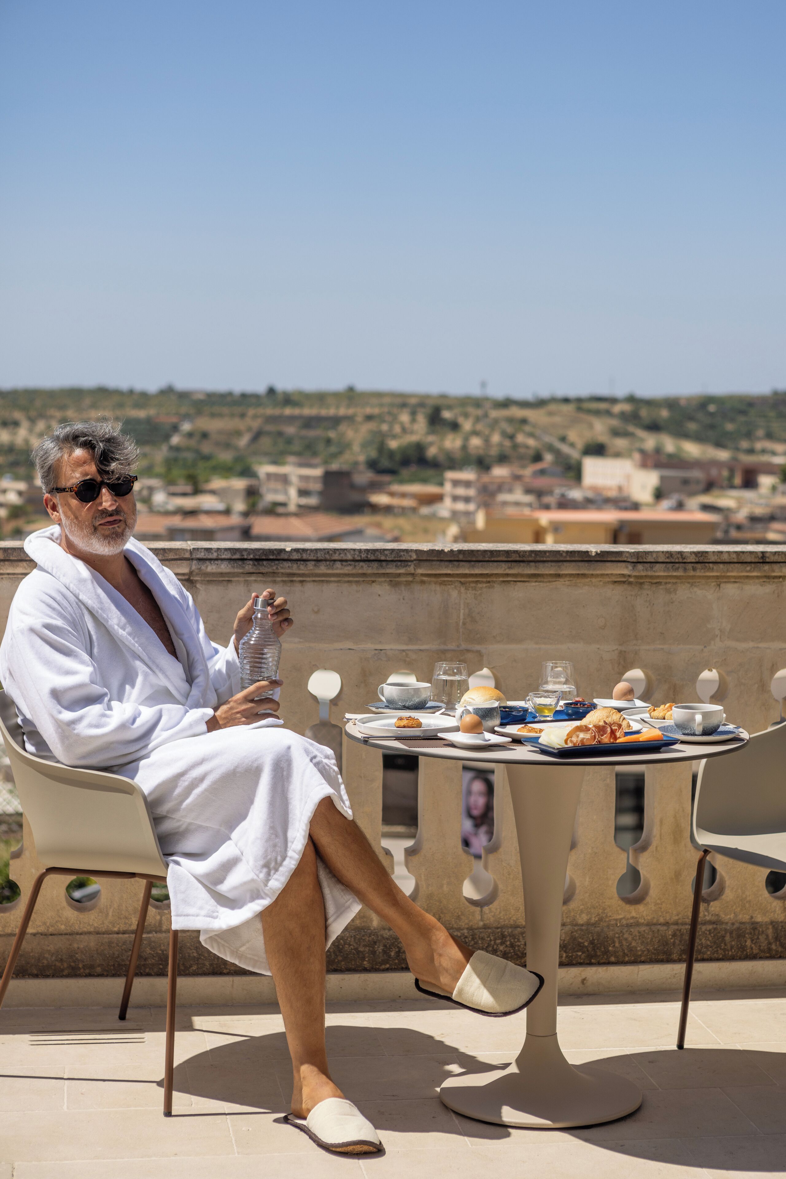 A man in a white robe sitting at a table with a view of a cityscape.