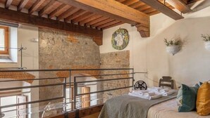 A bedroom with a stone wall, wooden beams, a bed with pillows, a small table with towels, and a window.