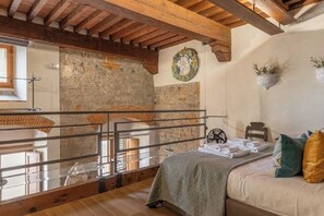 Un dormitorio con pared de piedra, vigas de madera, una cama con almohadas, una mesita con toallas y una ventana.