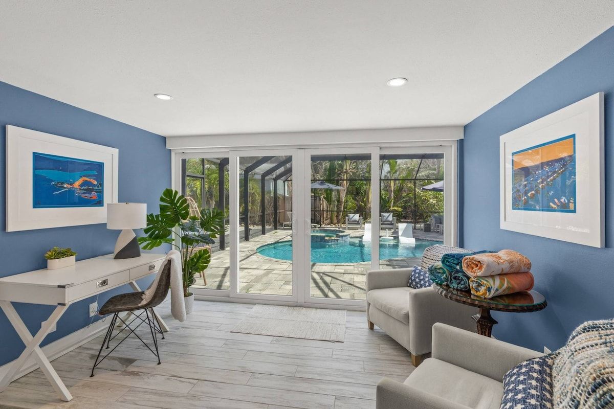 A room with a desk, a chair, a sofa, and a view of a swimming pool through large glass doors.