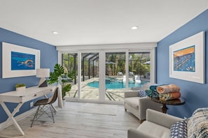 A room with a desk, a chair, a sofa, and a view of a swimming pool through large glass doors.