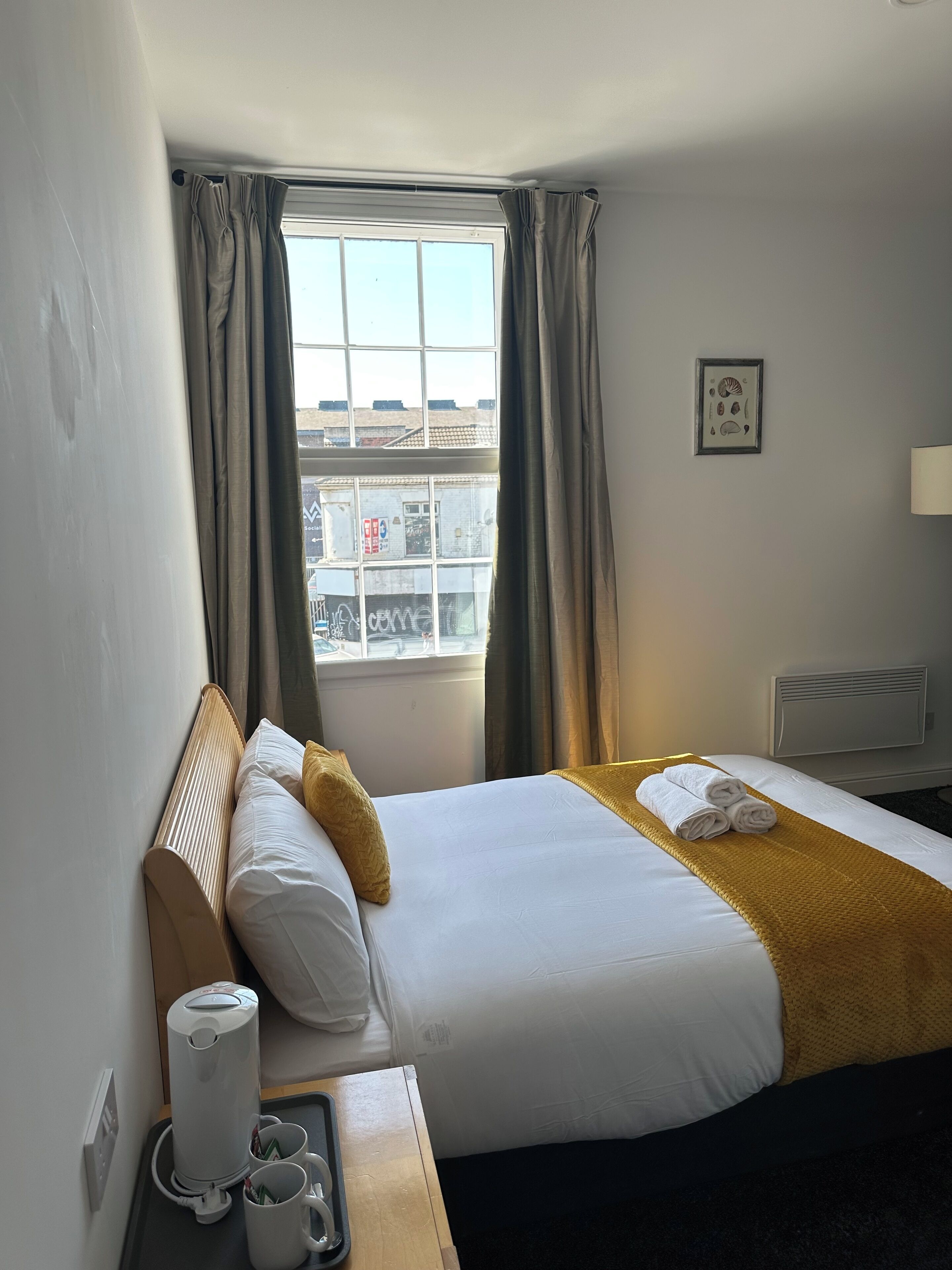 A hotel room with a bed, a bedside table with a coffee maker and mugs, a window with curtains, and a wall-mounted picture.