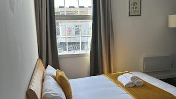 A hotel room with a bed, a bedside table with a coffee maker and mugs, a window with curtains, and a wall-mounted picture.