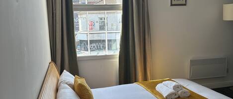 Habitación de hotel con cama, mesita de noche con cafetera y tazas, ventana con cortinas, y un cuadro colgado en la pared.