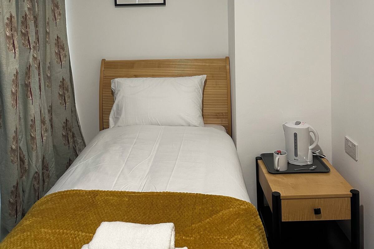 Hotellihuone, jossa on sänky, puinen pääty, seinällä kehystetty taulu, yöpöytä, jossa on vedenkeitin, ja sinapinvärinen heittopeitto.