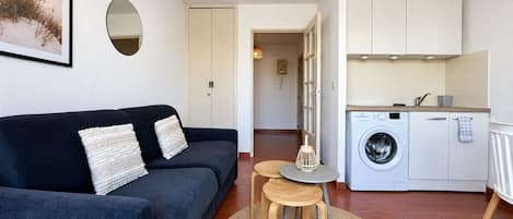 Una sala de estar con un sofá oscuro, un espejo redondo y una cocina con lavadora.