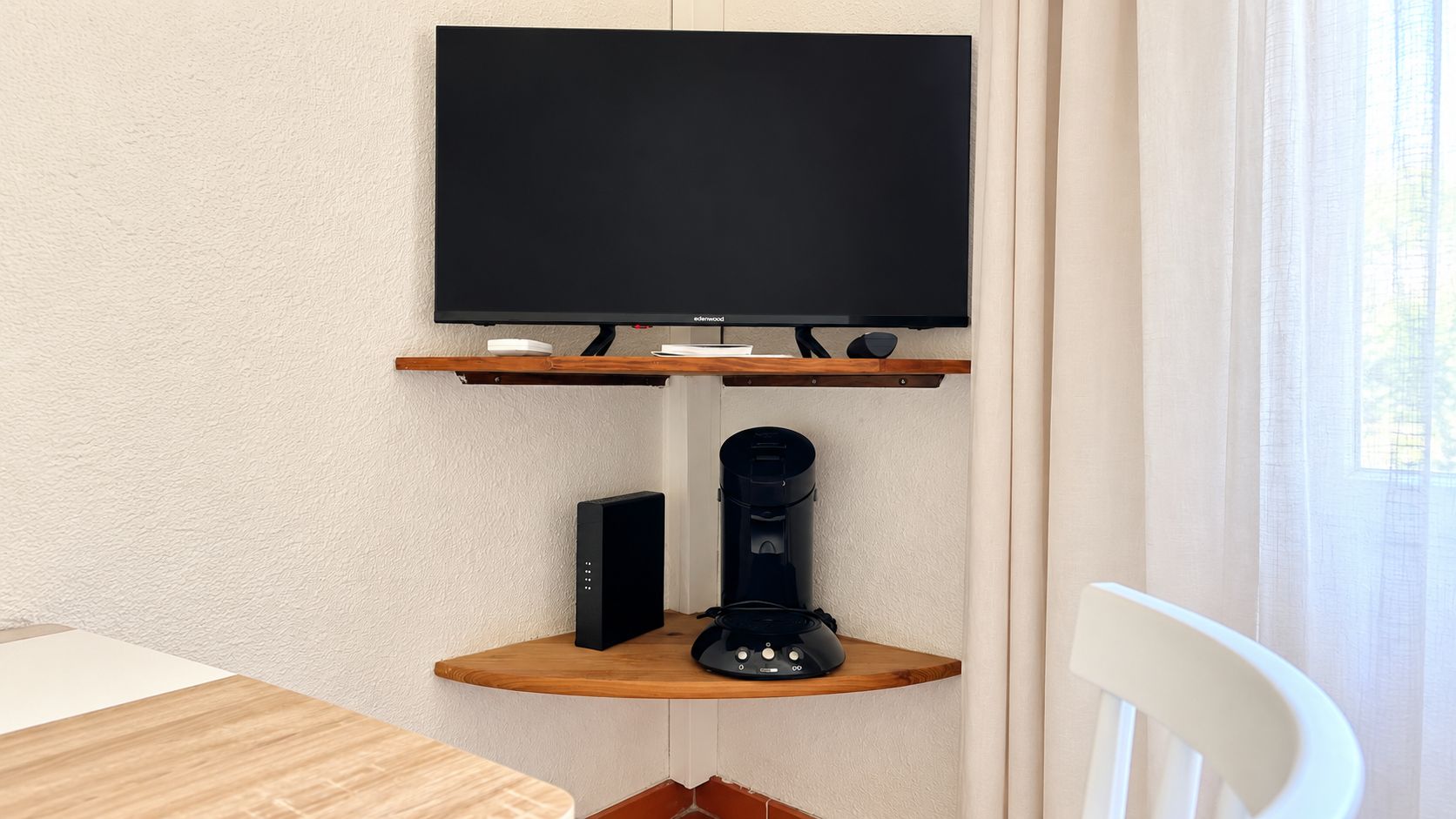 A flat-screen TV mounted on a wall above a wooden shelf with a coffee maker and a speaker.