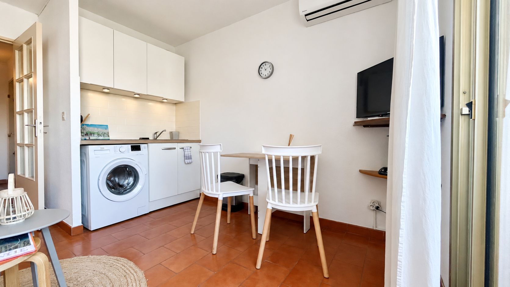 A compact kitchen with a washing machine, a dining table with chairs, and a TV on the wall.