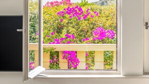 Ein Zimmer mit geöffnetem Fenster, das einen Balkon mit leuchtenden Blumen und Blick auf das Meer zeigt.