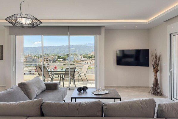 A modern living room with a sofa, a dining table with chairs, a TV mounted on the wall, and a view of the city through large windows.