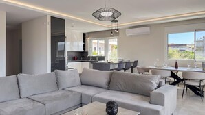 A modern living room with a grey sectional sofa, a coffee table, and a dining area with a table and chairs. There is a kitchen in the background with white cabinets and a large window. - Ardians Home, Be on Holidays (Chania)