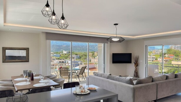 A modern living room with a sofa, dining table, and large windows offering a city view. - Ardians Home, Be on Holidays (Chania)
