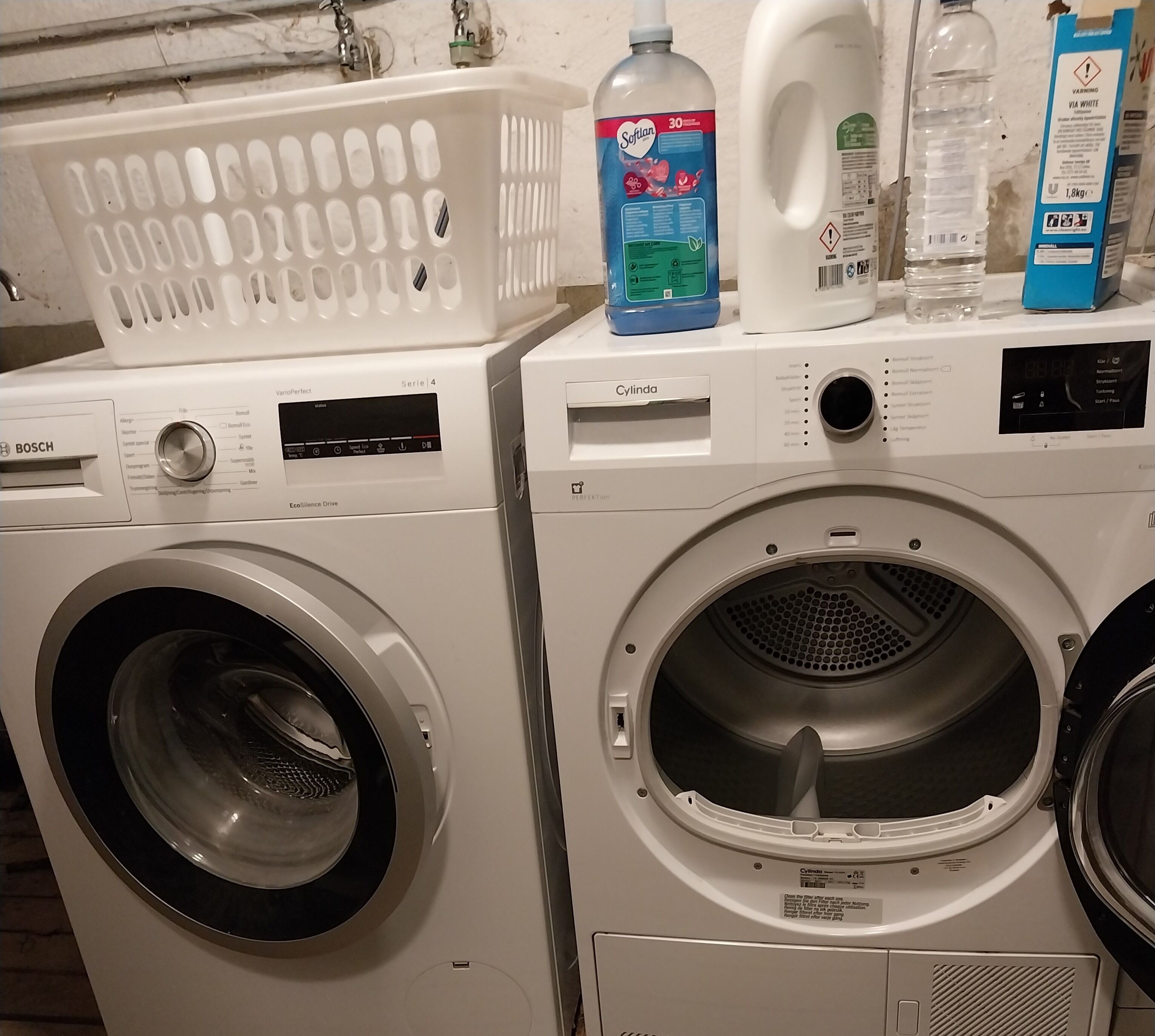 Dos lavadoras de carga frontal con paneles de control, una cesta de ropa blanca y una botella de detergente Softlan.