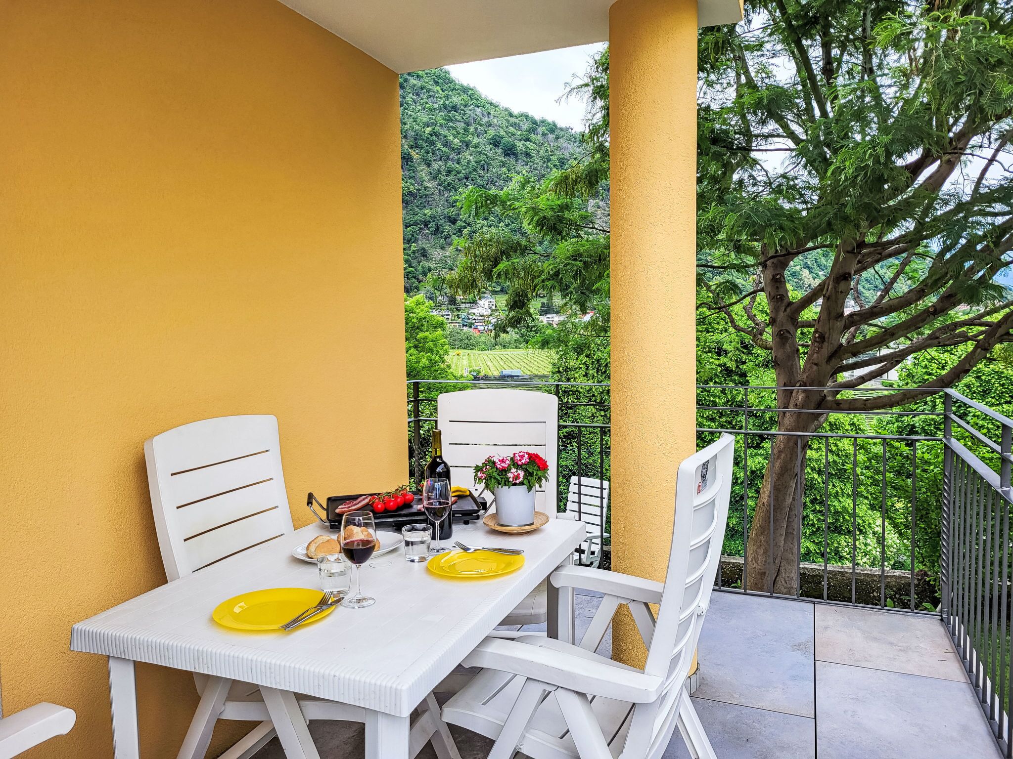Un balcon avec une table dressée pour deux personnes, des chaises blanches et une assiette jaune.