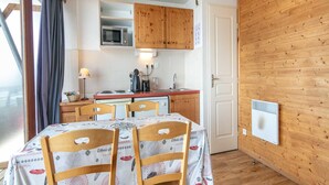 A compact kitchen with wooden cabinets, a microwave, and a stove. There is a dining table with chairs, a window with curtains, and a wall-mounted heater.
