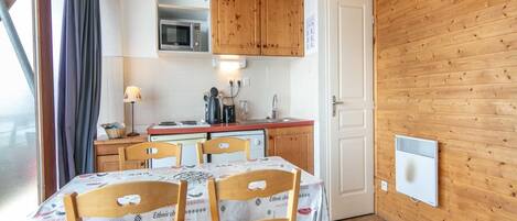 A compact kitchen with wooden cabinets, a microwave, and a stove. There is a dining table with chairs, a window with curtains, and a wall-mounted heater.
