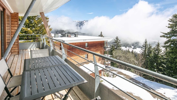 A balcony with a wooden deck and metal railing overlooking a snowy landscape and a mountain range.