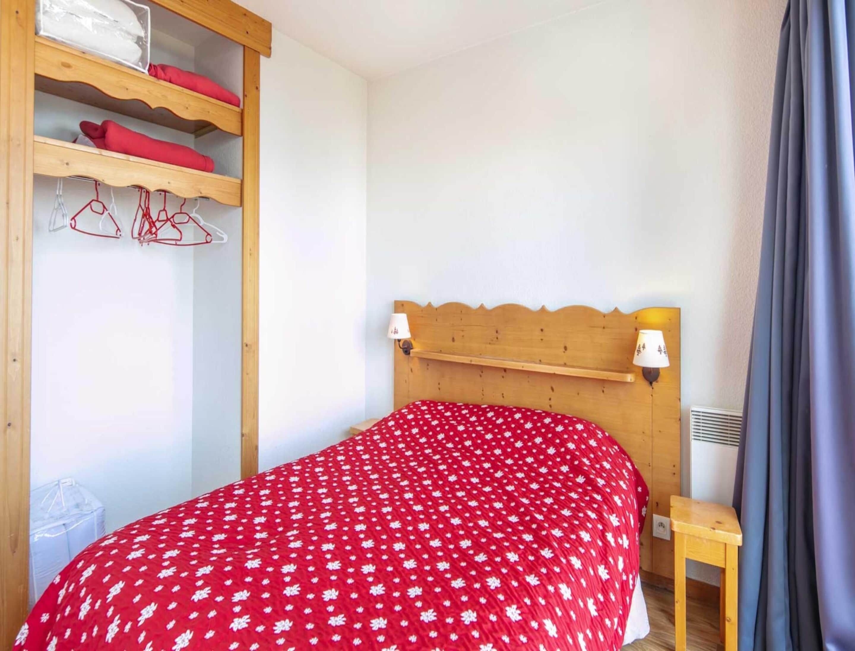 Ein Schlafzimmer mit einem Holzkopfteil, einer roten blumigen Tagesdecke, einem Holzschrank mit Kleidung und einem kleinen Nachttisch.