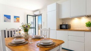 A dining area with a wooden table set for four, white chairs, and a vase with flowers. A kitchen with white cabinets and a microwave is visible in the background. - Apartment Gemma by Interhome (Valledoria)