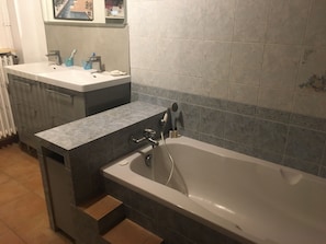 Et badeværelse med badekar, vask og underskab. Der er trin op til badekarret, og et håndklædestativ er synligt.