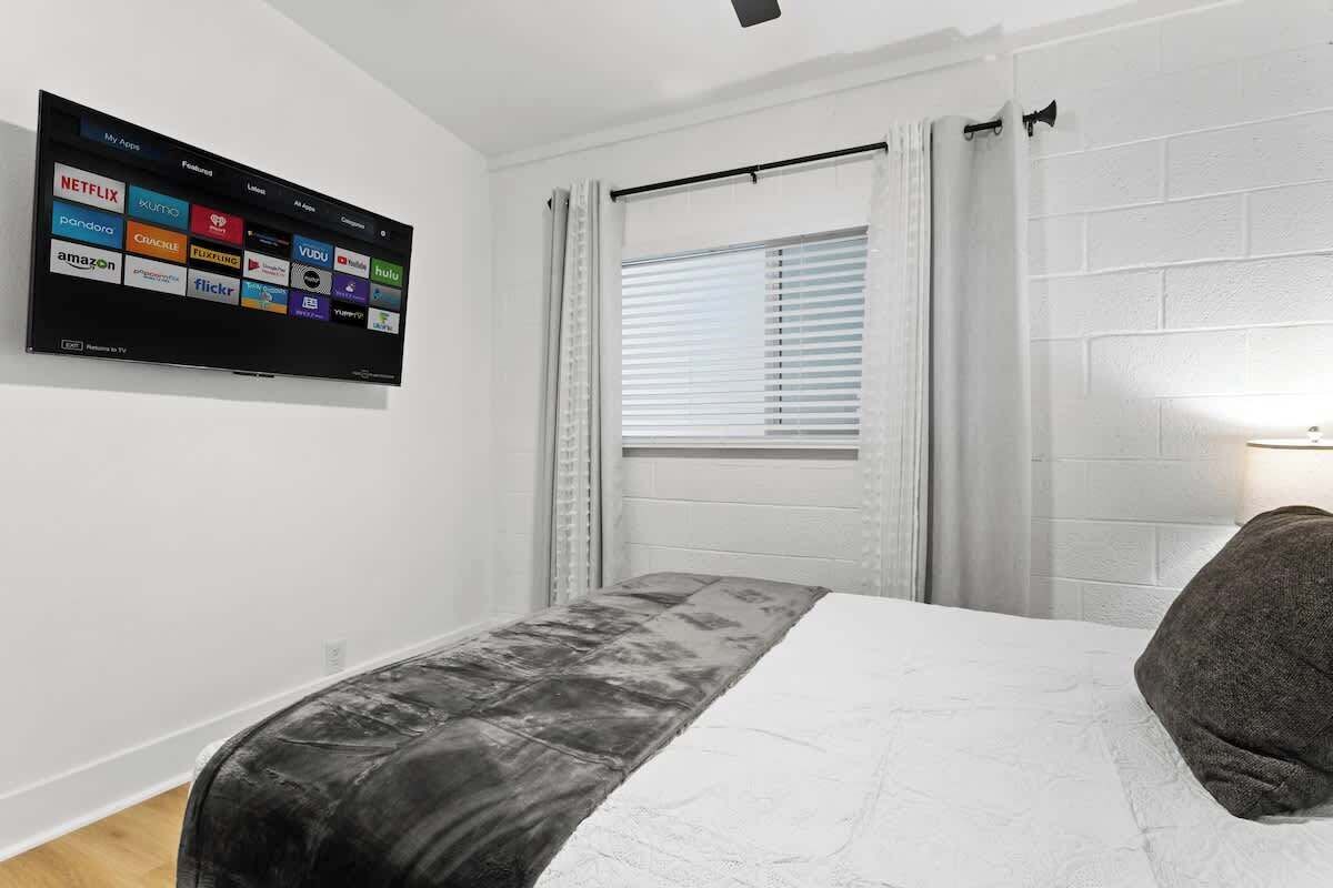 Ein Hotelzimmer mit Bett, einem an der Wand befestigten Flachbildfernseher, der verschiedene Streaming-Dienste anzeigt, einem Fenster mit Jalousien und einer Nachttischlampe.