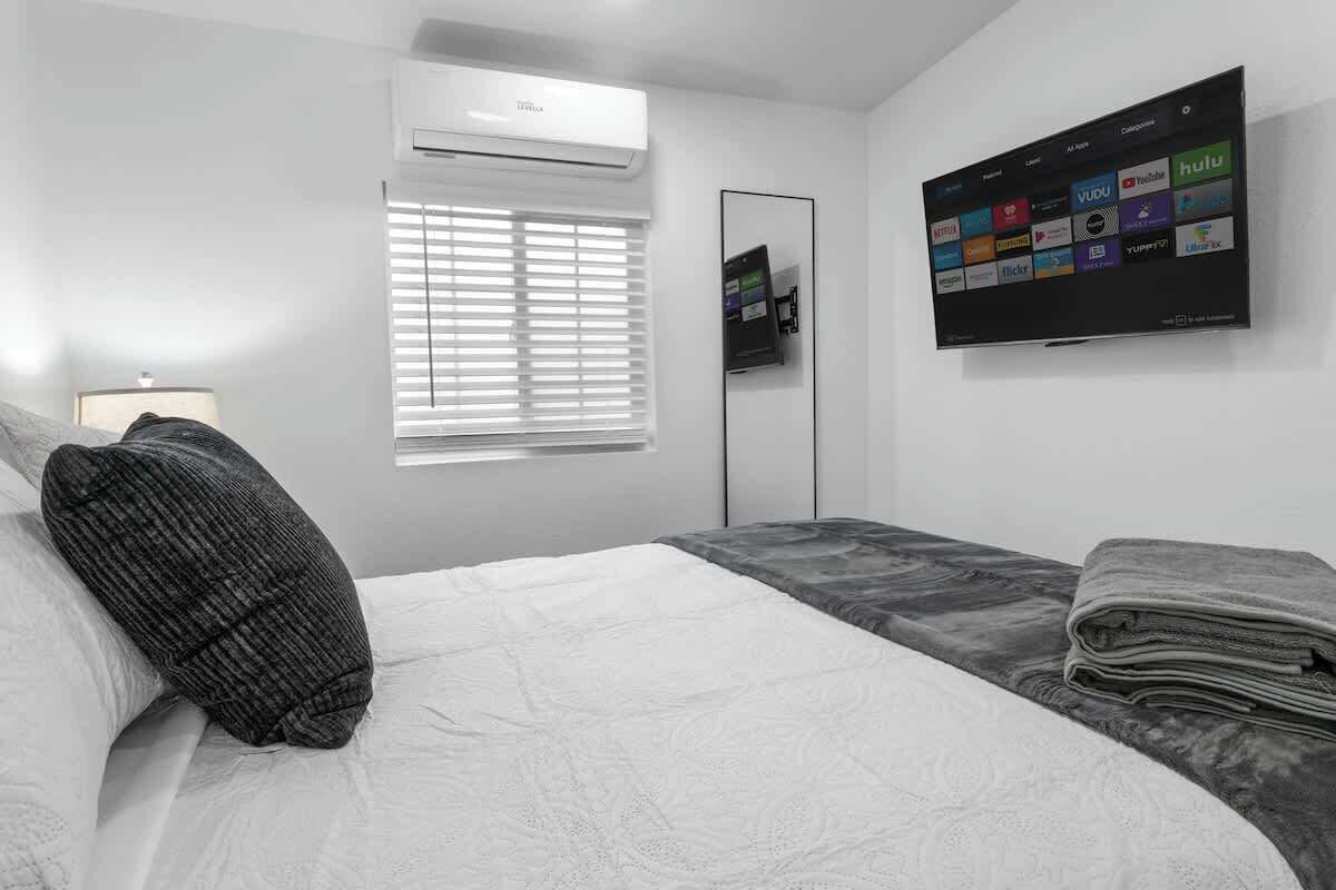 Una habitación de hotel moderna con una cama grande, un televisor de pantalla plana montado en la pared que muestra varios servicios de streaming, una ventana con persianas y un aire acondicionado instalado en la pared.