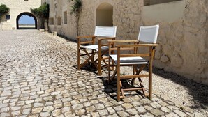 Une cour pavée de pierres, avec deux chaises en bois et une table, un mur de pierre et une porte en arc donnant sur un bâtiment dont le toit est rouge.