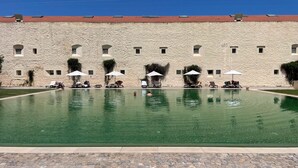 Une piscine entourée de chaises longues et de parasols, elle-même bordée d’un bâtiment en pierre doté de nombreuses fenêtres.