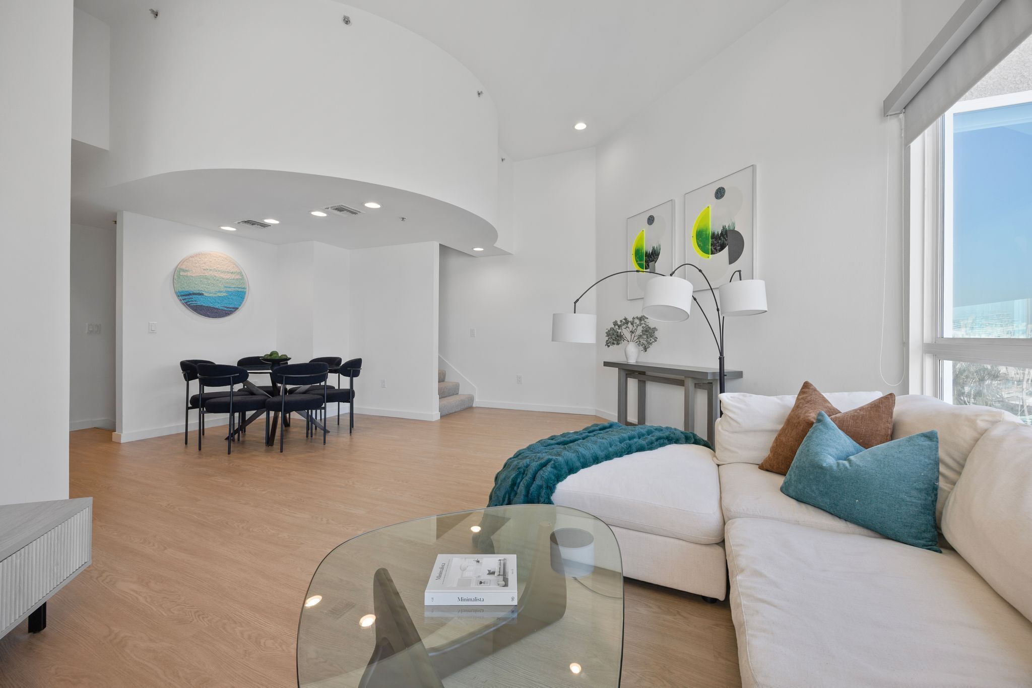 A modern living room with a glass coffee table, a sofa with cushions, a dining area with chairs, and artwork on the walls.