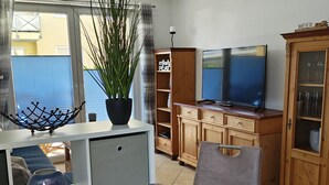 A living room with a TV, wooden cabinets, a plant, and a dining area with a blue table and chairs. - Ostsee-Urlaub-Zempin is perfect for your time-out, right by the coastal forest. (Zempin)