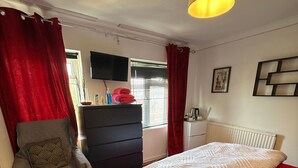 A bedroom with a bed, a chair, a dresser, a television, and a window with curtains. - Room-king Size - Ensuite Room (London)