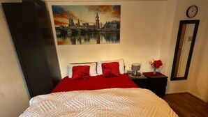 A hotel room with a large bed, a painting of a cityscape, a clock, and a mirror on the wall. - Room-king Size - Ensuite Room (London)