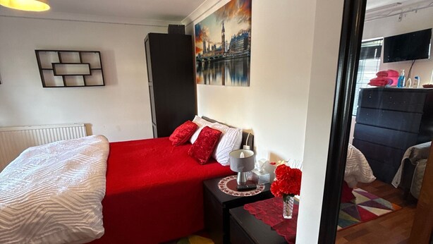 A bedroom with a red bed, a bedside table with a lamp, a clock on the wall, and a painting of a cityscape. - Room-king Size - Ensuite Room (London)