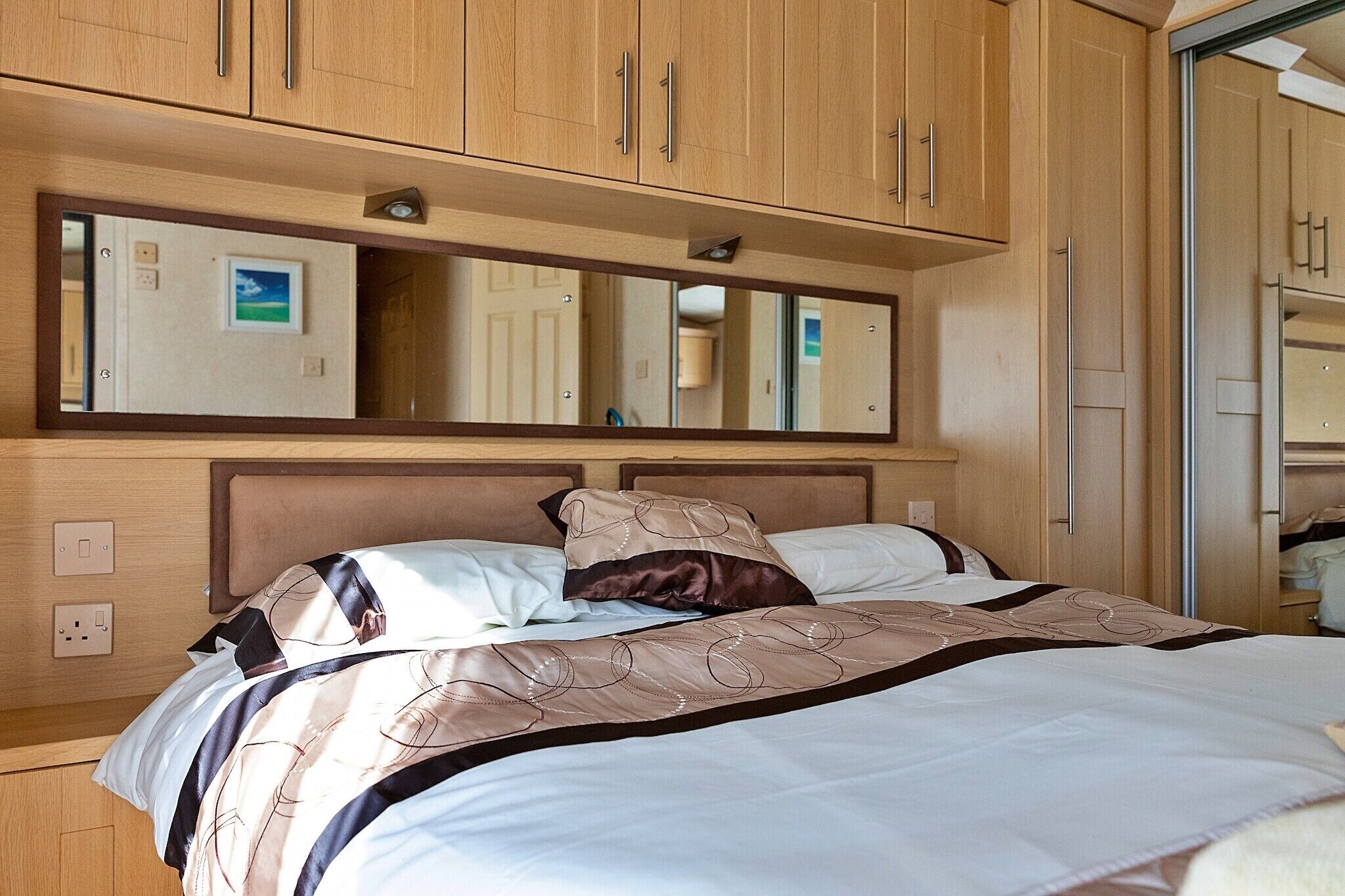 A bedroom with a bed, a large mirror, wooden cabinets, and a wall-mounted light fixture.