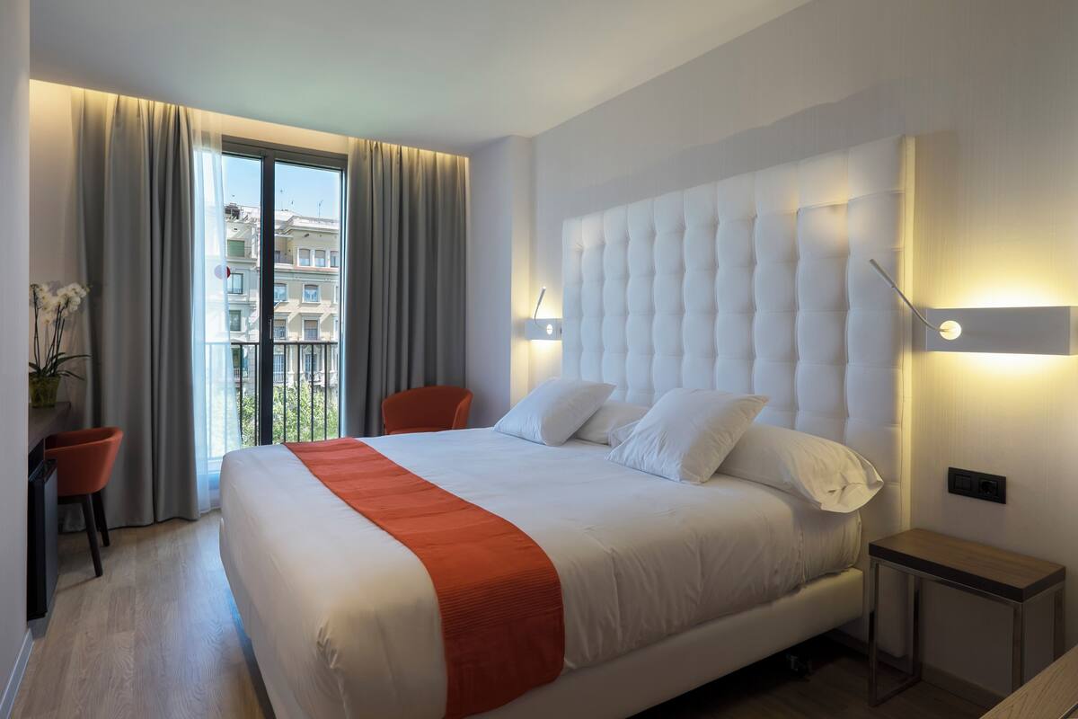 A hotel room with a large bed, a nightstand, a chair, and a view of the city through the window.