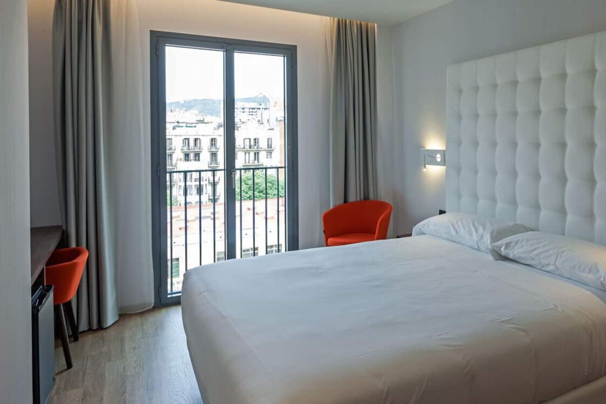 A hotel room with a large bed, a view through a sliding door, a red chair, and a modern headboard with wall-mounted lights.