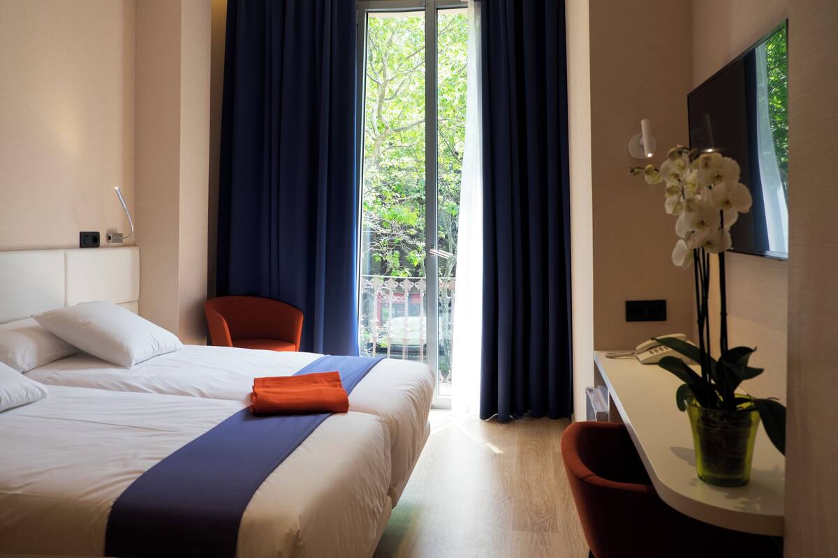 A hotel room with a bed, a desk, a chair, a potted plant, and a view of the outdoors.
