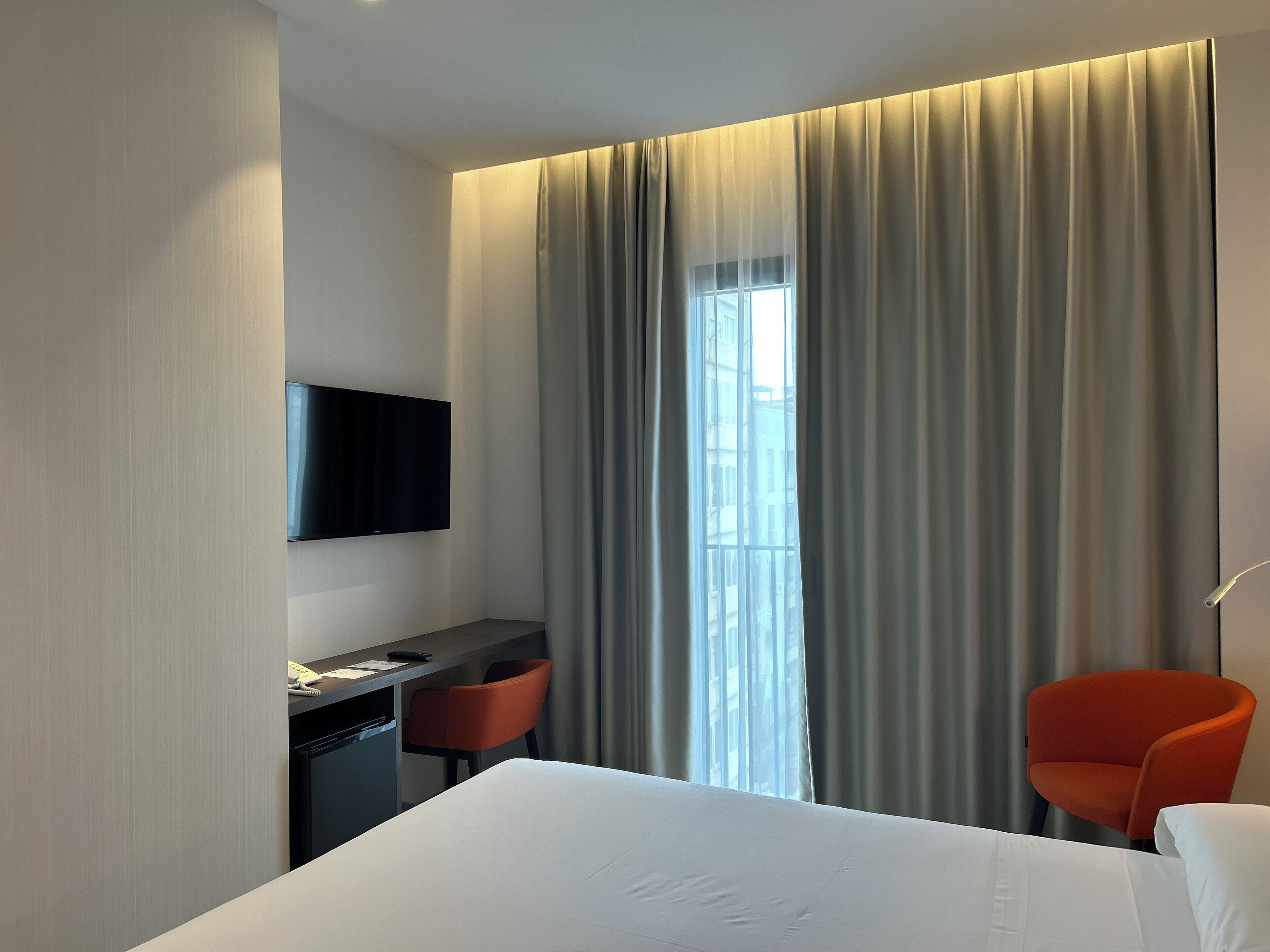 Une chambre d’hôtel comprenant un lit, un bureau avec une chaise, un téléviseur fixé au mur et une fenêtre avec des rideaux.