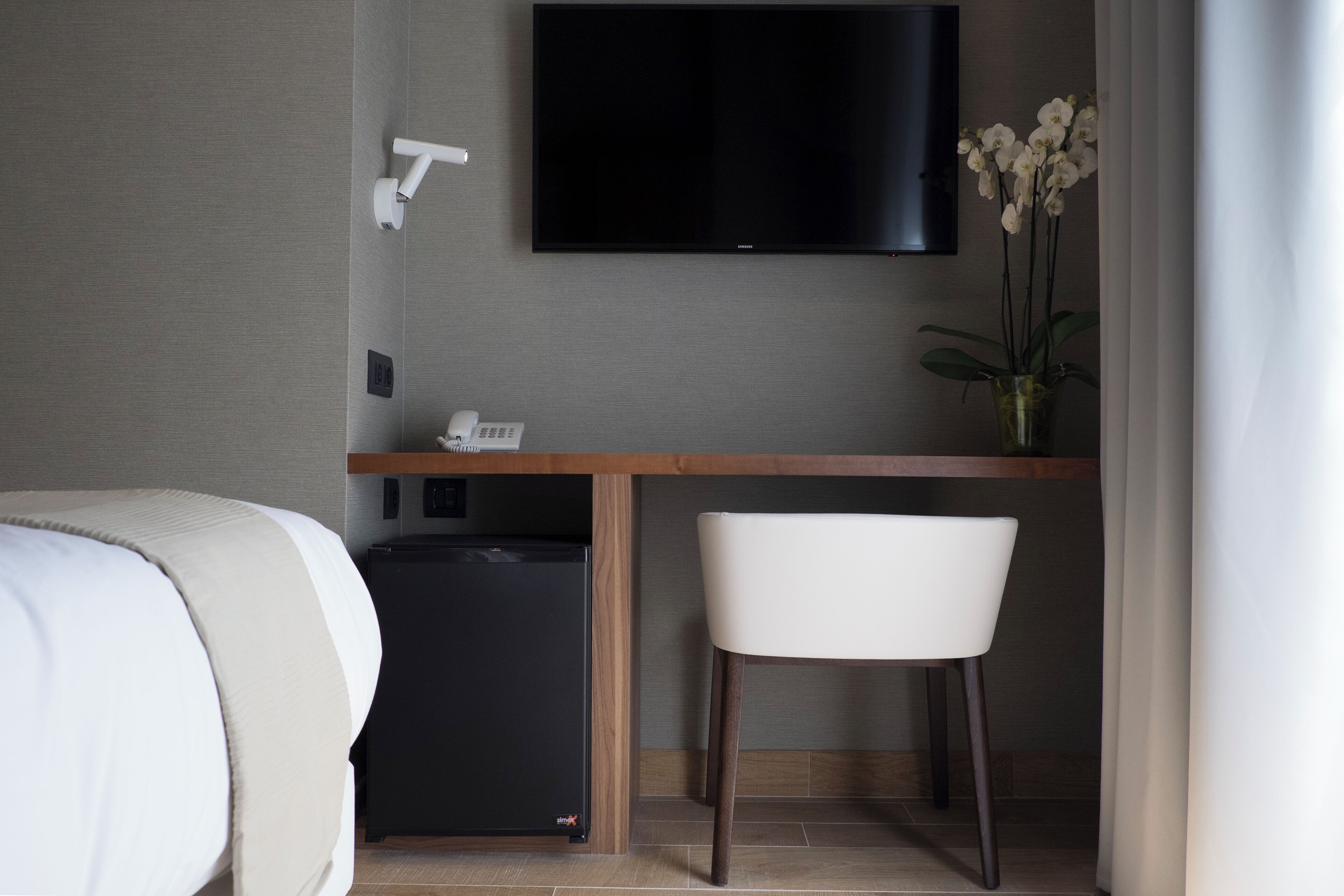 Habitación de hotel con cama, escritorio con silla, televisor, minibar y un florero con flores.