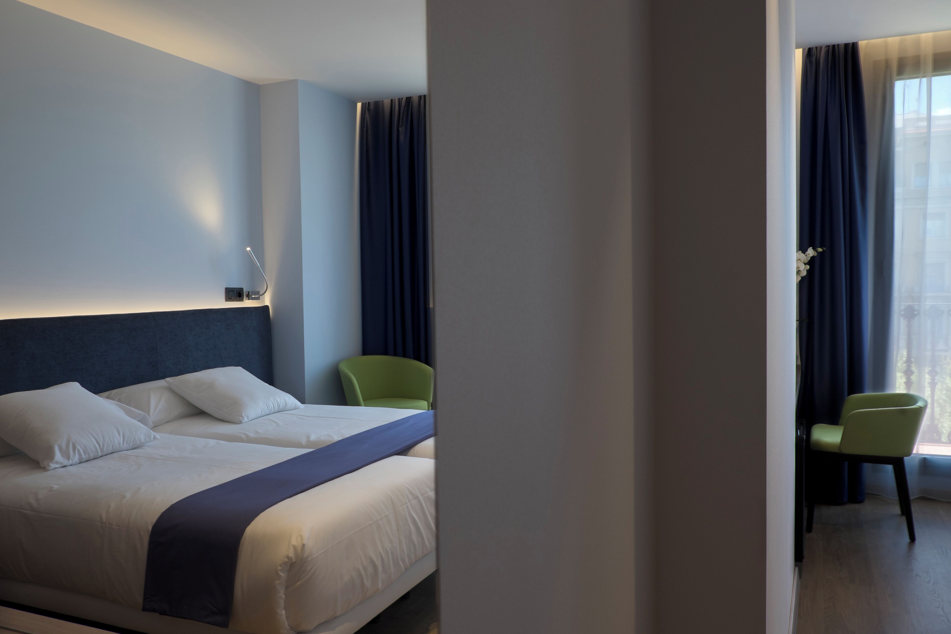 A hotel room with a bed, two chairs, a nightstand, and a window with curtains.