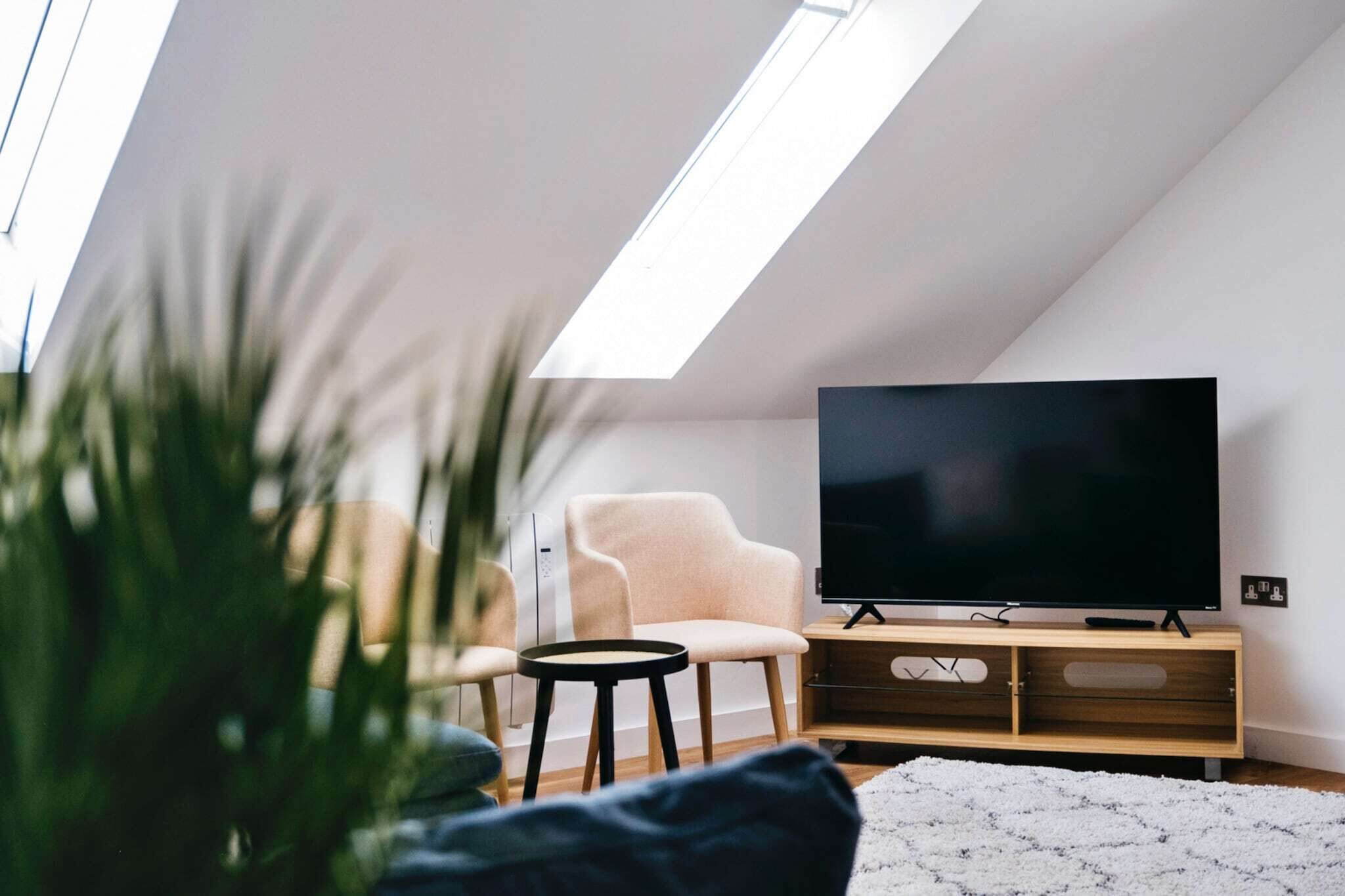 Un saló modern amb un televisor de pantalla plana sobre un suport de fusta, un butac armchair de color clar, una taula rodona petita i una catifes blanca i esponjosa.