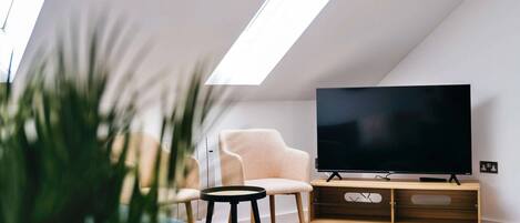 Un saló modern amb un televisor de pantalla plana sobre un suport de fusta, un butac armchair de color clar, una taula rodona petita i una catifes blanca i esponjosa.