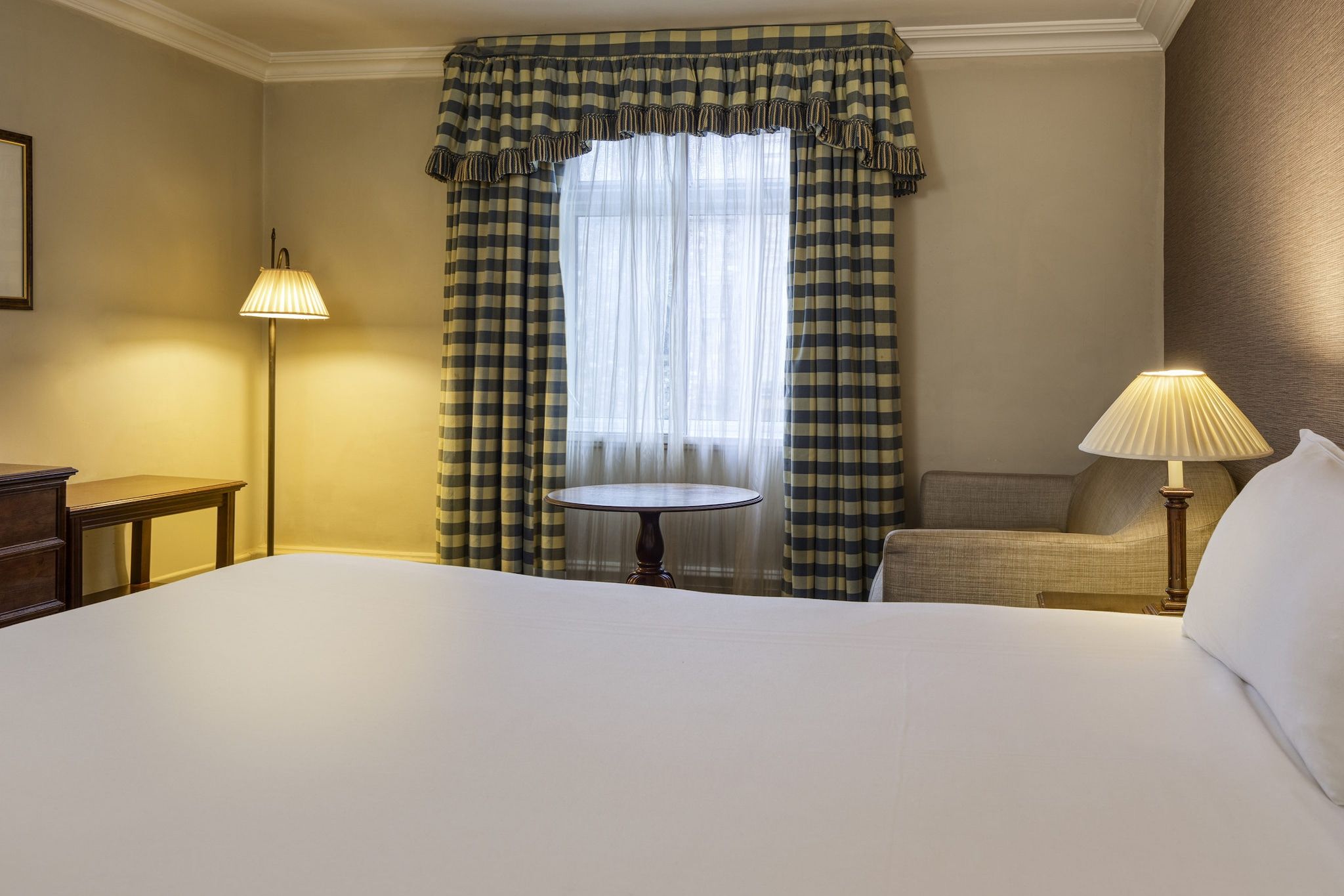 Hotellihuone, jossa on suuri sänky, pyöreä pöytä, kaksi yöpöytävalaisinta ja ikkuna, jossa on ruutukuviolliset verhot.