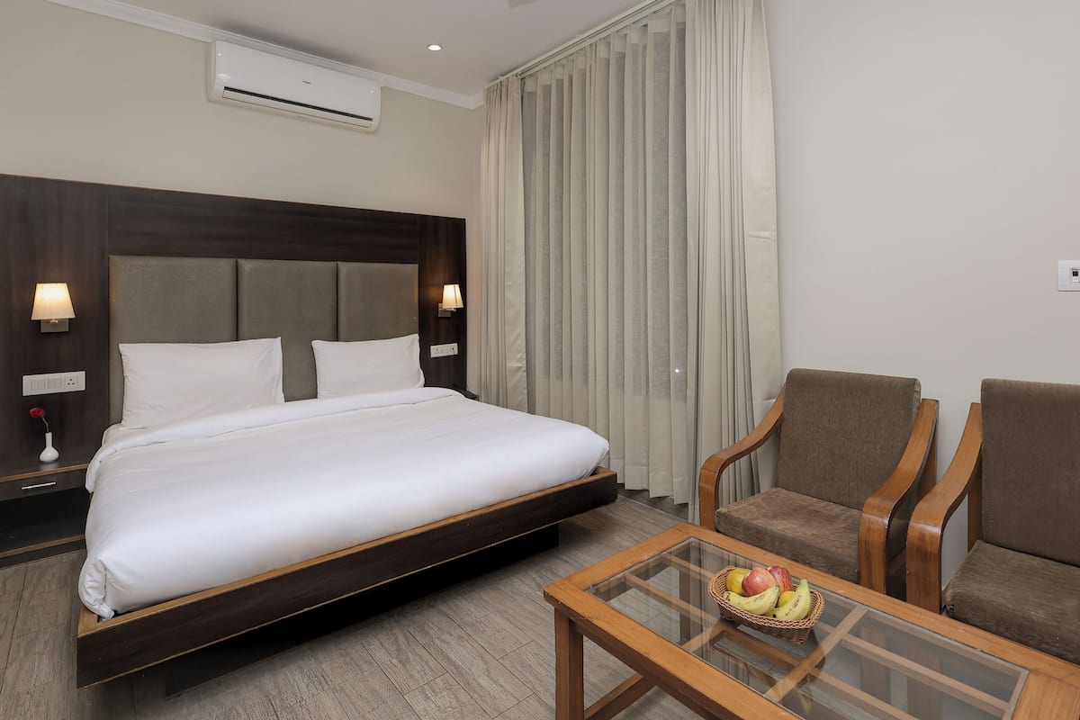 A hotel room with a large bed, two bedside lamps, a wooden chair, a glass table with fruit, and a window with curtains.