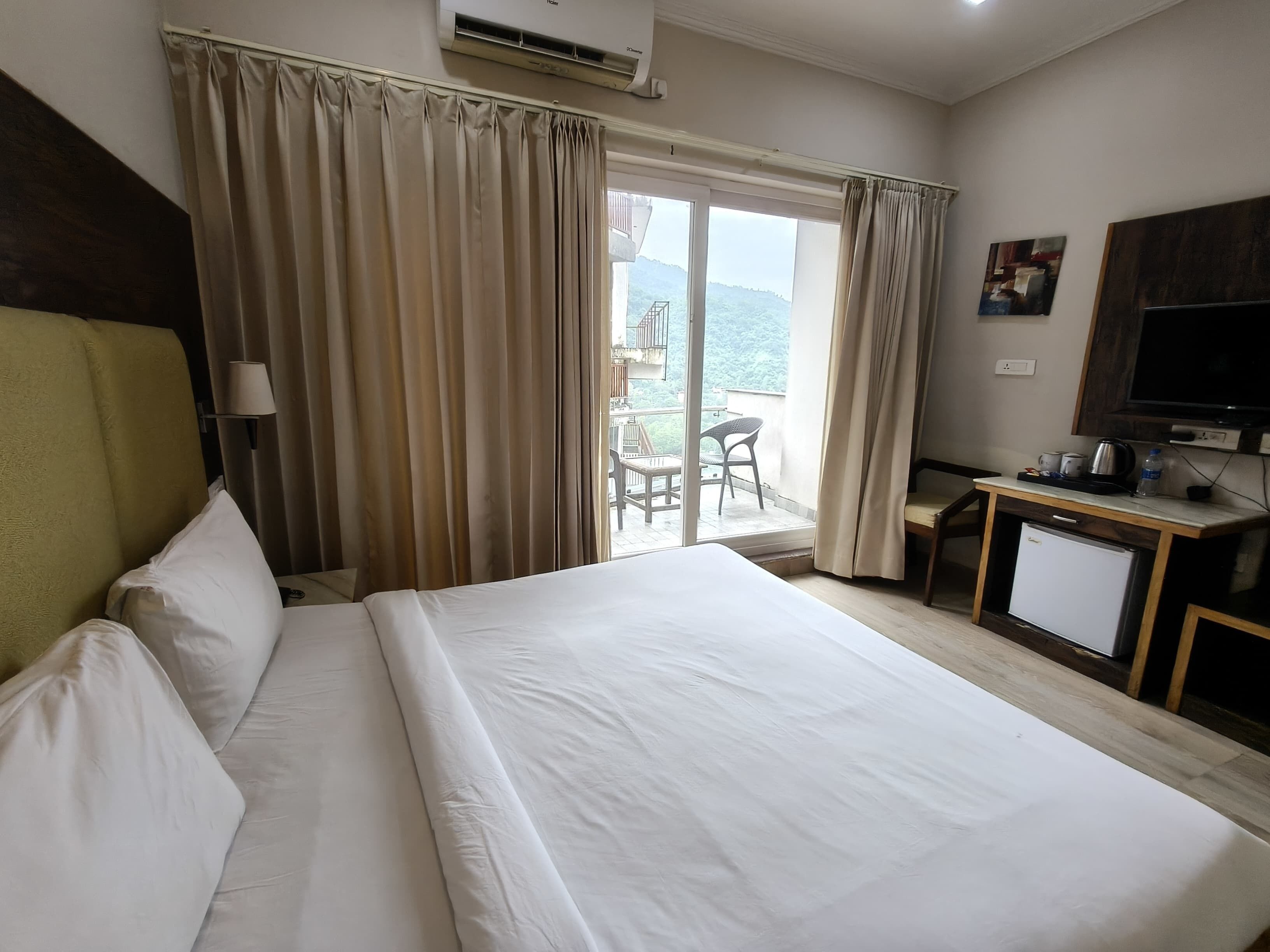 A hotel room with a bed, a desk with a television, and a view of a balcony with outdoor furniture.